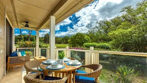 A covered outdoor patio with wicker furniture and a table set for dining, overlooking a lush garden and a body of water. - Grand Champions 29 1 Bedroom Condo (Kihei)