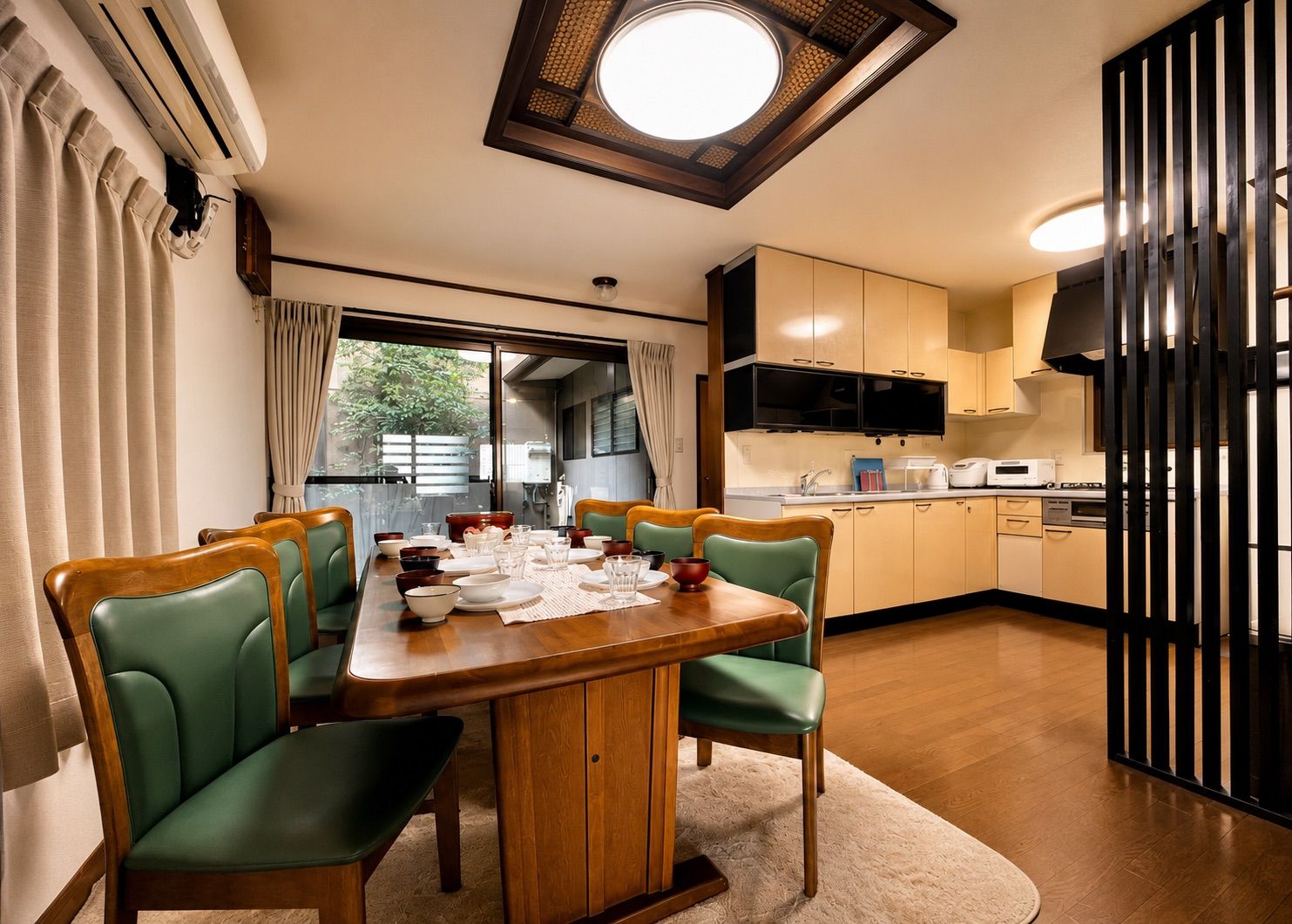 A dining area with a wooden table set for four, surrounded by green chairs, a kitchen with modern appliances, and a large window providing natural light.