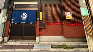 Un edificio de dos pisos con fachada oscura, techo de tejas rojas y una unidad de aire acondicionado grande montada en la pared. Hay dos puertas de madera grandes con una reja de metal, y un toldo azul con un logotipo sobre la entrada.