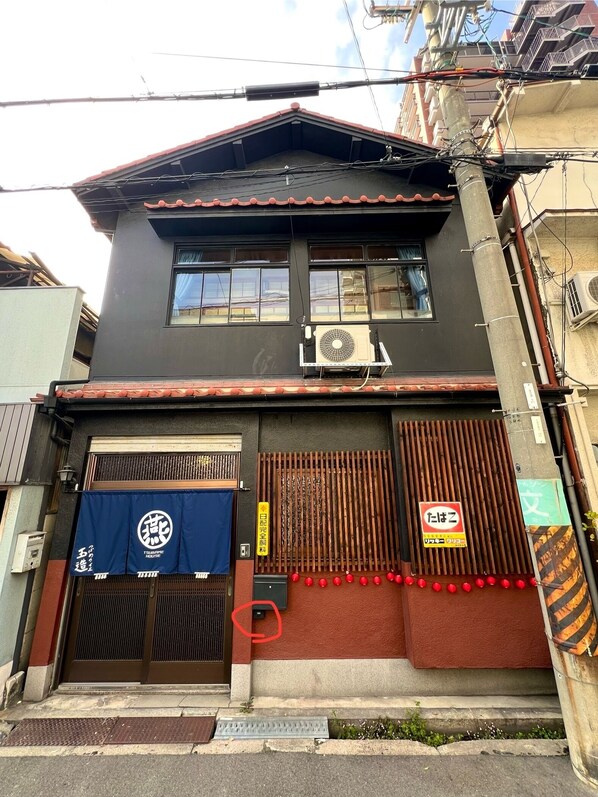 A two-story building with a dark facade, a red-tiled roof, and a large air conditioning unit mounted on the wall. There are two large wooden doors with a metal grill, and a blue awning with a logo above the entrance. - [Tsubame no Ie Tamatsukuri] 1-minute walk from Tamatsukuri Station | Accommodates up to 8 guests | 100 ㎡ | Shinsaibashi, Namba, Umeda, Nara, Osaka Castle | Excellent access (大阪市)