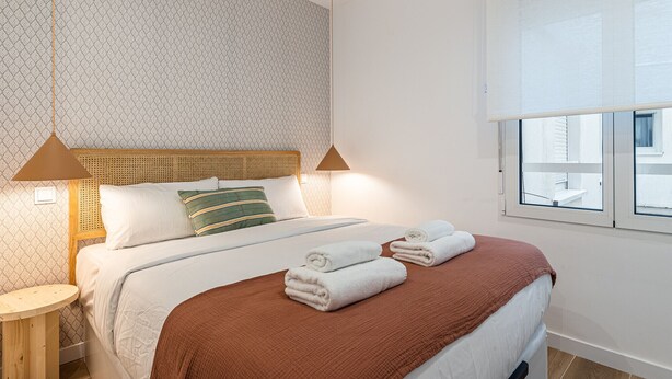 A neatly made bed with a brown blanket, two pillows, and rolled towels on the side. There is a window with blinds, a bedside table with a lamp, and a patterned wall.