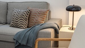 Een grijze bank met gekleurde kussens en een blauwe deken, een houten stoel en een zwarte vloerlamp.