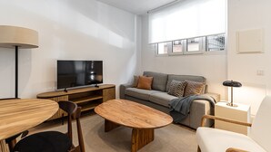 Een moderne woonkamer met een flatscreen televisie, houten meubels, een bank met opvallende kussens en een groot raam met zonwering.