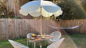 Een houten tafel met een witte parasol, omringd door witte stoelen, op een gazon met een hangmat op de achtergrond.