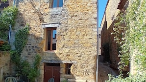 A stone building with a small window and a door, surrounded by greenery and a table with chairs.