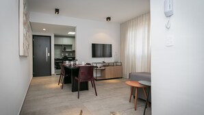A modern living room with a dining area, a kitchen in the background, and a flat-screen TV mounted on the wall. - Moderno apartamento 2 quartos (Belo Horizonte)