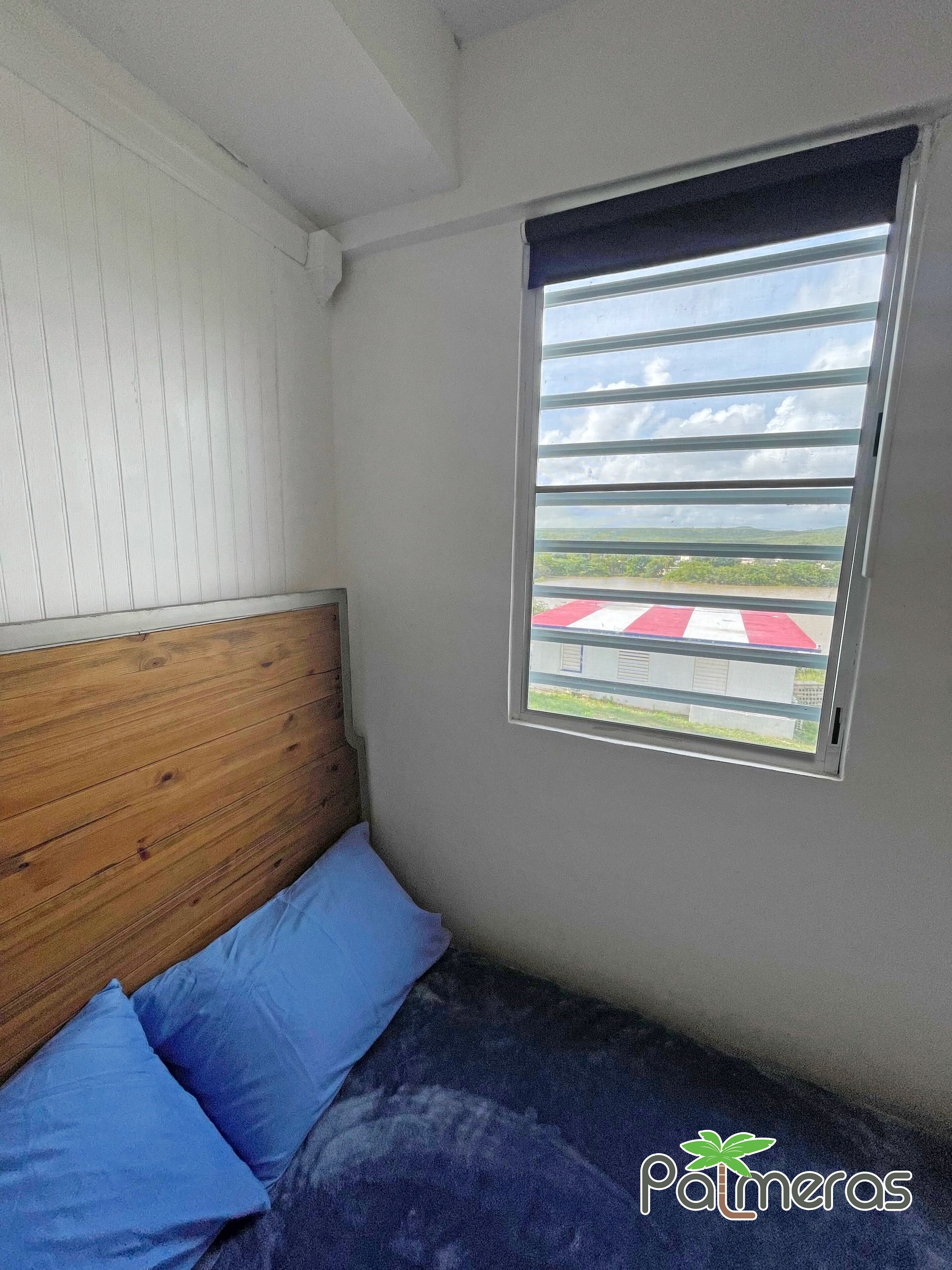 Ein Schlafzimmer mit einem hölzernen Kopfteil, einem Einzelbett mit blauer Bettwäsche und einem Fenster mit horizontalen Rollläden.