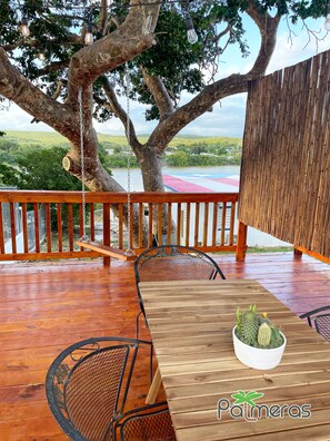 A wooden deck with a table and chairs, a hanging light fixture, and a view of a river and greenery. - Beach Apartment with Terrace in Playa Santa (Ensenada)
