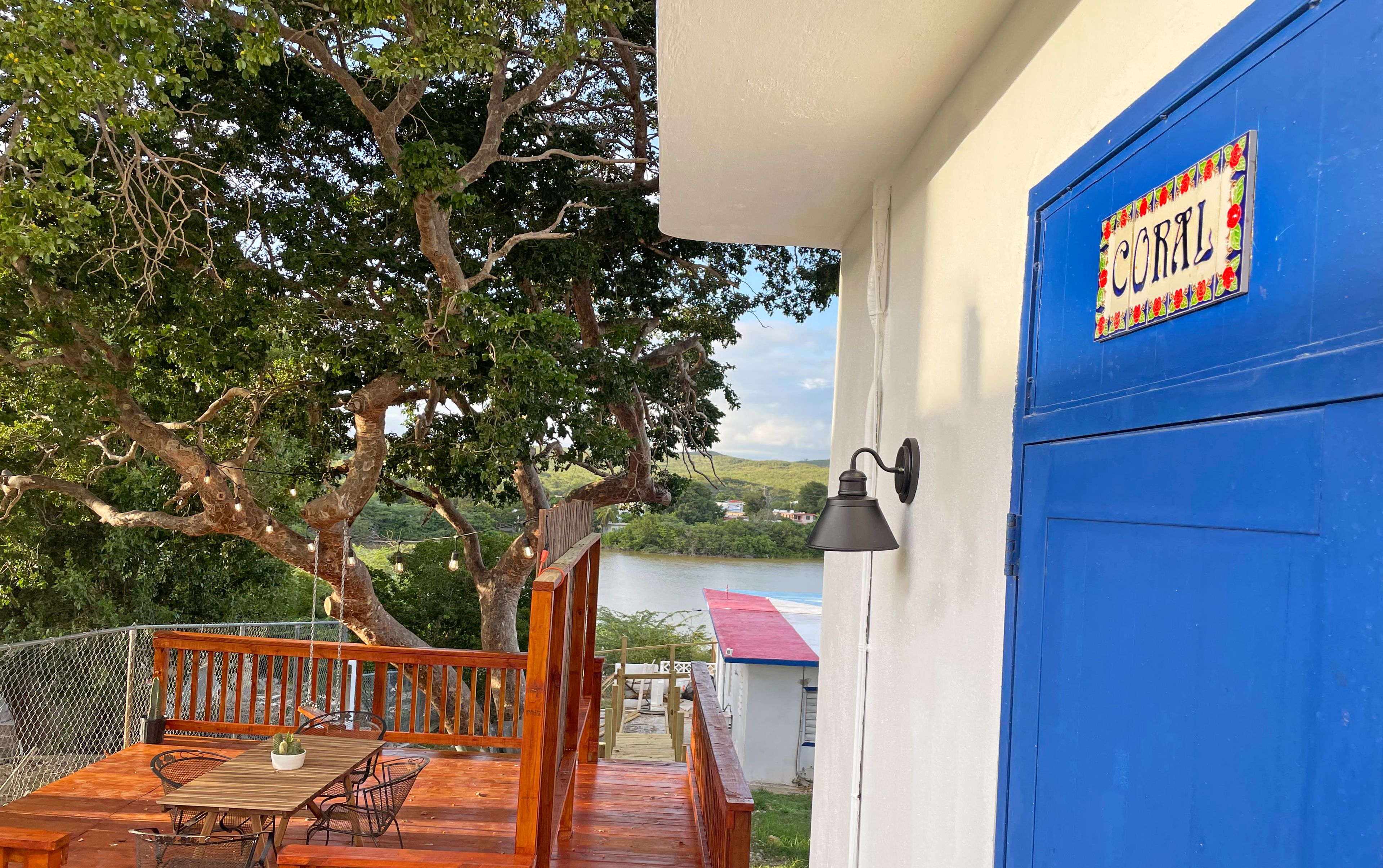 Una terraza de madera con una mesa y sillas, una puerta azul con placa, y vista a un lago.