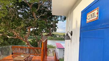 Una terraza de madera con una mesa y sillas, una puerta azul con placa, y vista a un lago.