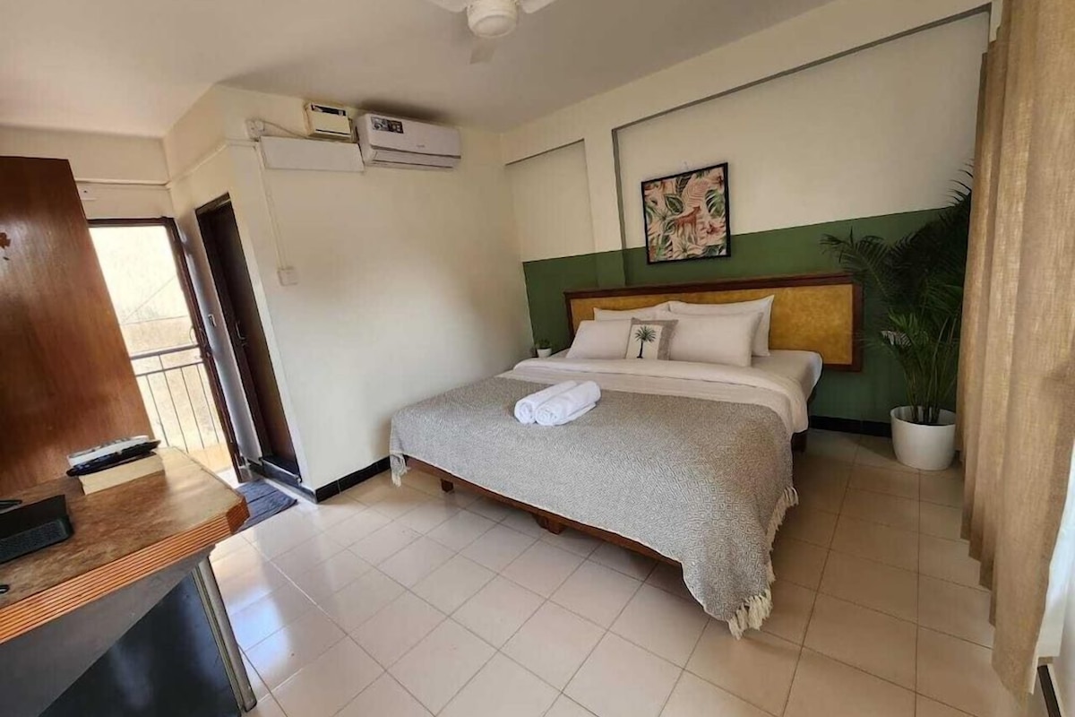A hotel room with a bed, a desk, a ceiling fan, an air conditioning unit, a potted plant, and a painting on the wall.