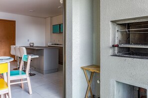 A modern kitchen with a central island, a dining area with a small table and chairs, and a view of a balcony with a grill. - Club Vista Mar Pertinho do Beto Carrero (Penha)