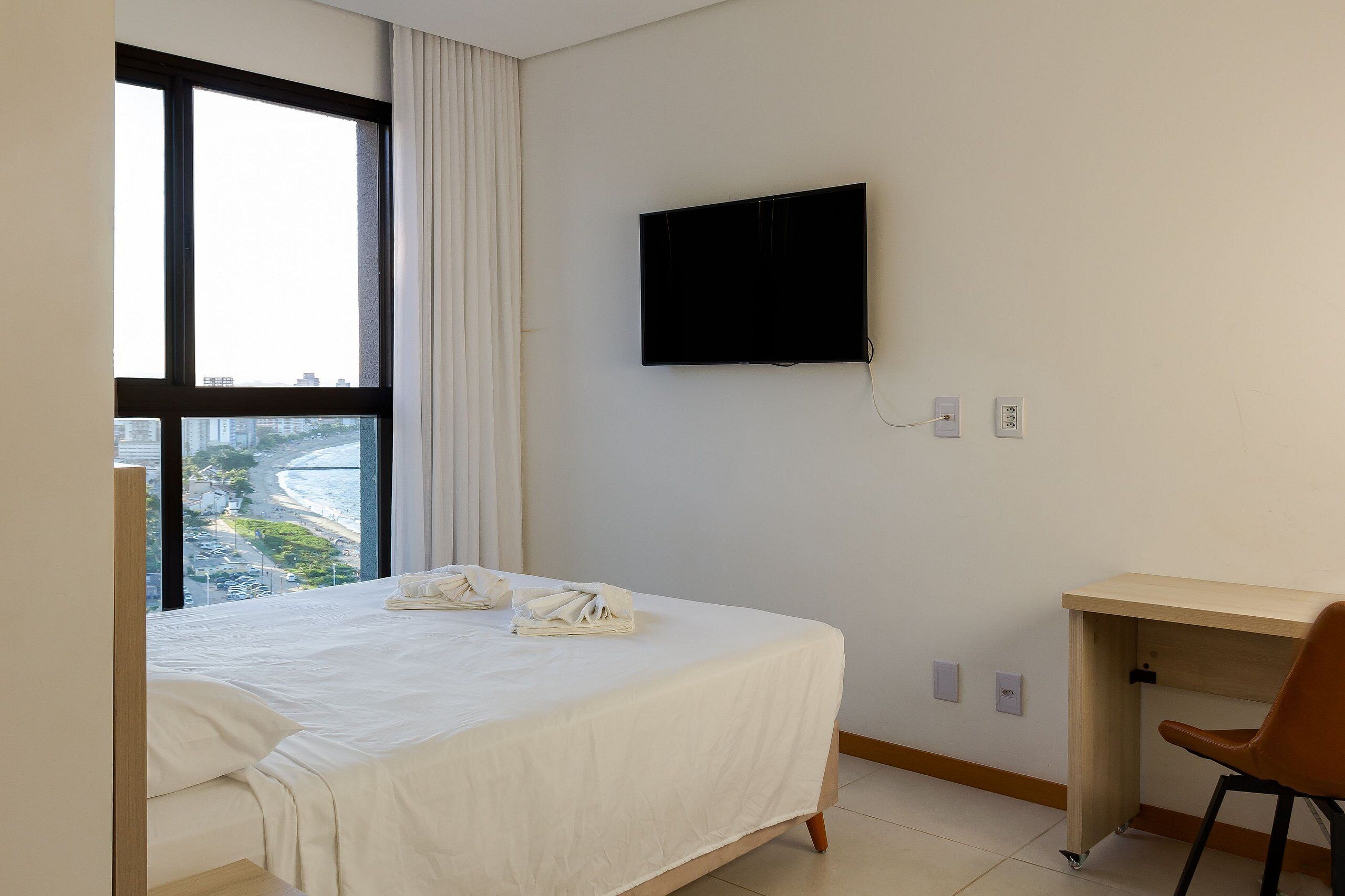 A hotel room with a bed, a desk, a chair, a TV, and a window with a view of a beach and buildings.