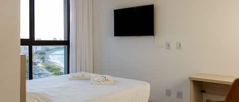Une chambre d’hôtel équipée d’un lit, d’un bureau, d’une chaise, d’une télévision et d’une fenêtre donnant sur une plage et des bâtiments.