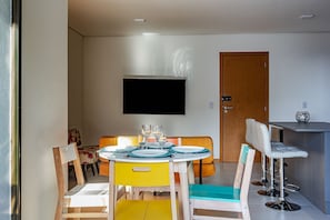 A dining area with a round table set for four, chairs with teal cushions, a wooden bench, and a television mounted on the wall. - Club Vista Mar Pertinho do Beto Carrero (Penha)