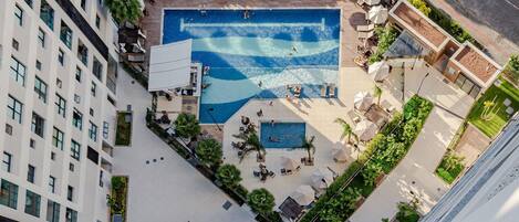 An aerial view of a hotel with a central swimming pool surrounded by sun loungers and umbrellas, with a building complex in the background.