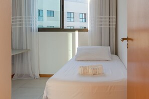A single bed with a pillow and a rolled towel, a window with curtains, and a view of a building outside. - Club Vista Mar Pertinho do Beto Carrero (Penha)