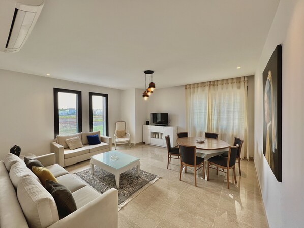 A modern living room with a sofa, coffee table, dining table, and chairs. There is a TV on a white cabinet, and a painting on the wall. - Residence Villa Jade Luxe Marsa (La Marsa)