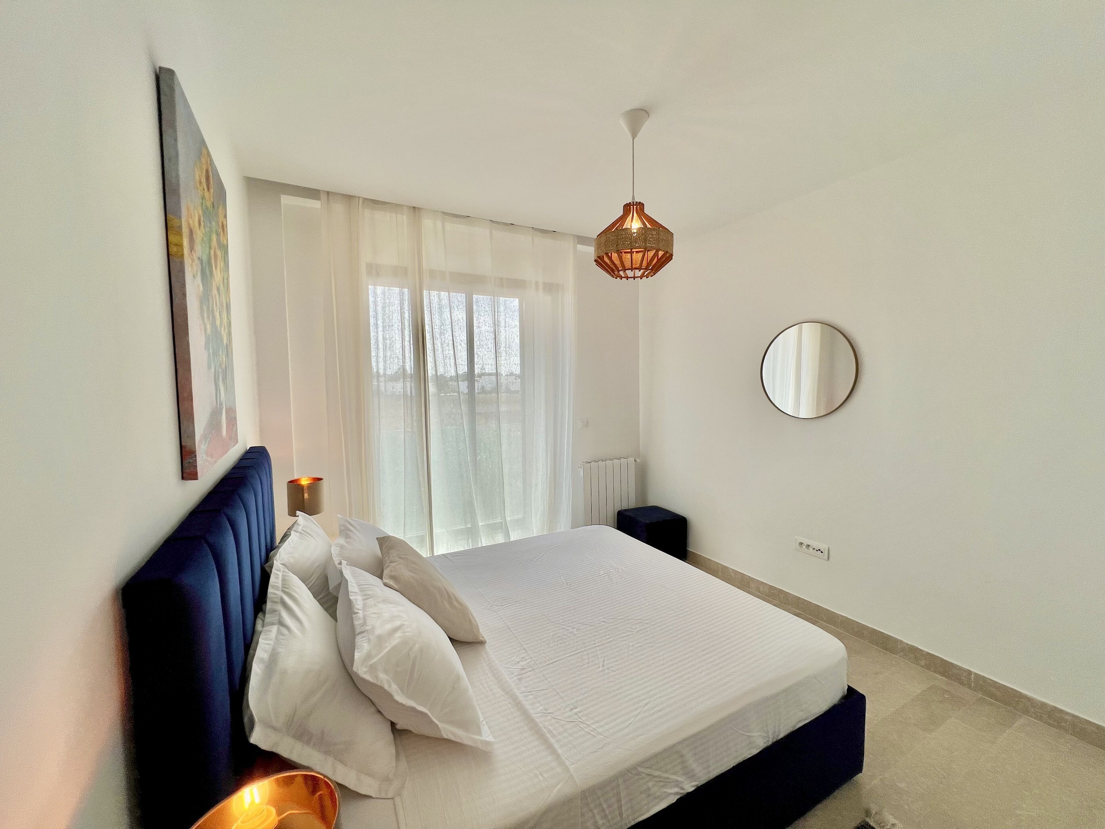 Una camera da letto con un letto, una lampada da comodino, un lampadario, una finestra con tende leggere e uno specchio fissato al muro.