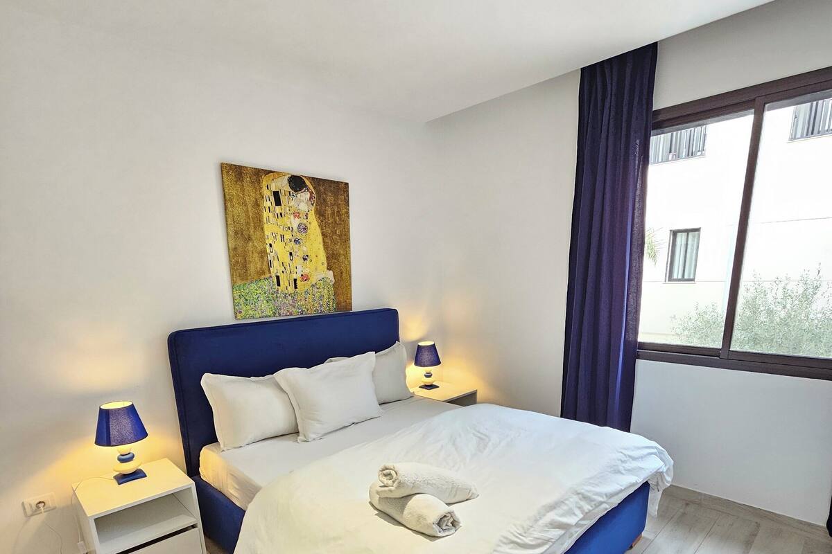 Ein modernes Schlafzimmer mit einem großen Bett, einem Nachttisch mit Lampe, einem Gemälde an der Wand und einem Fenster mit Vorhängen.