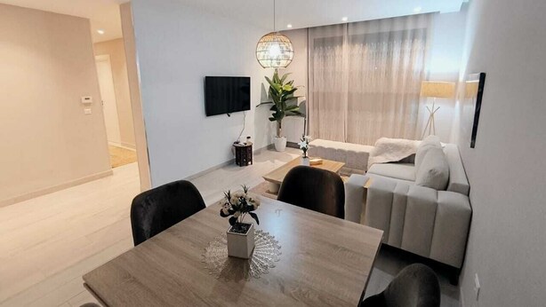 Een moderne woonkamer met een eethoek, een grijze bank, een aan de muur bevestigde televisie en een potplant.