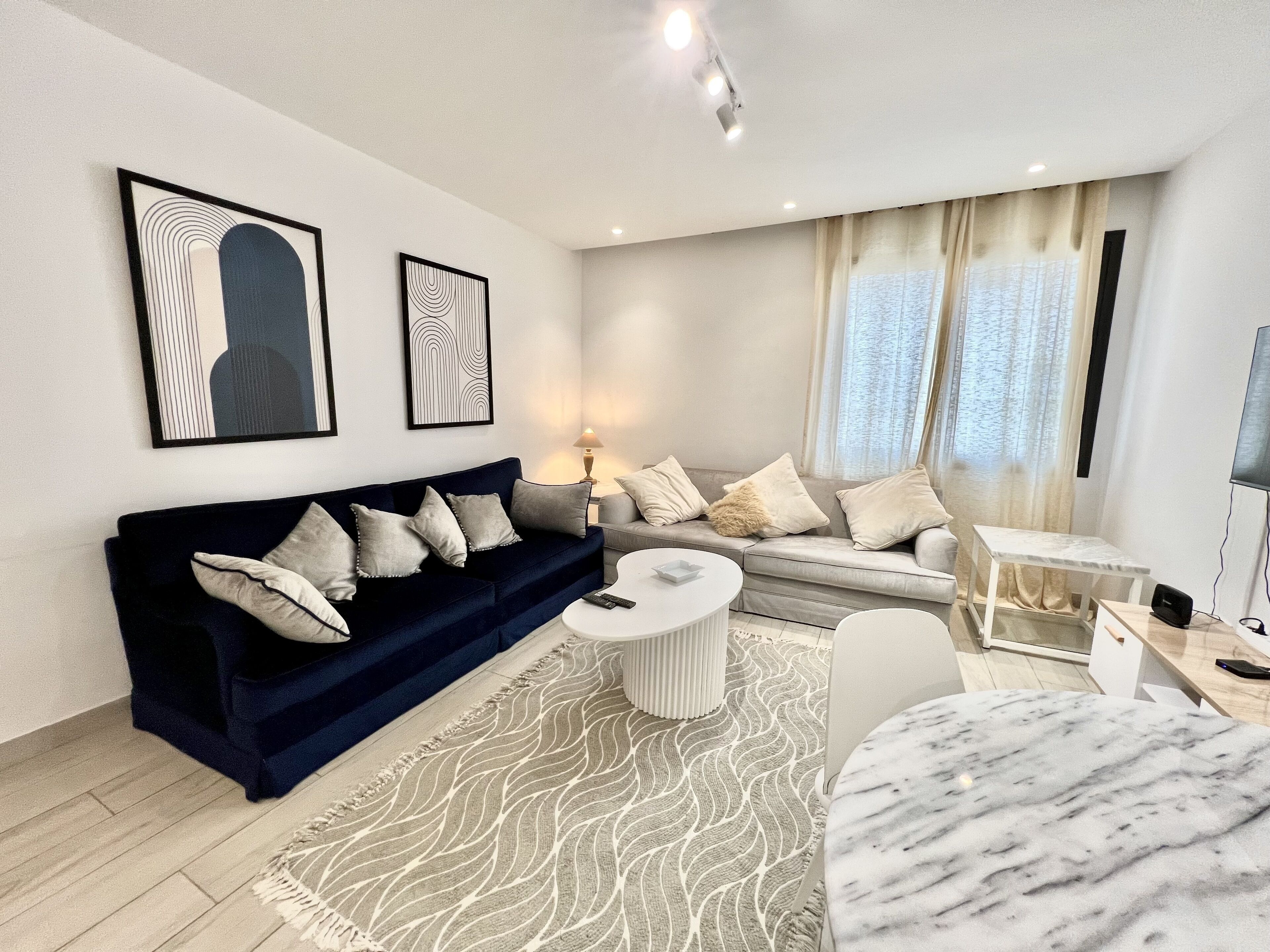 Un salon moderne avec un canapé bleu foncé, une table basse blanche et un tapis à motifs. Deux œuvres d’art encadrées ornent le mur, et une fenêtre est agrémentée de rideaux.