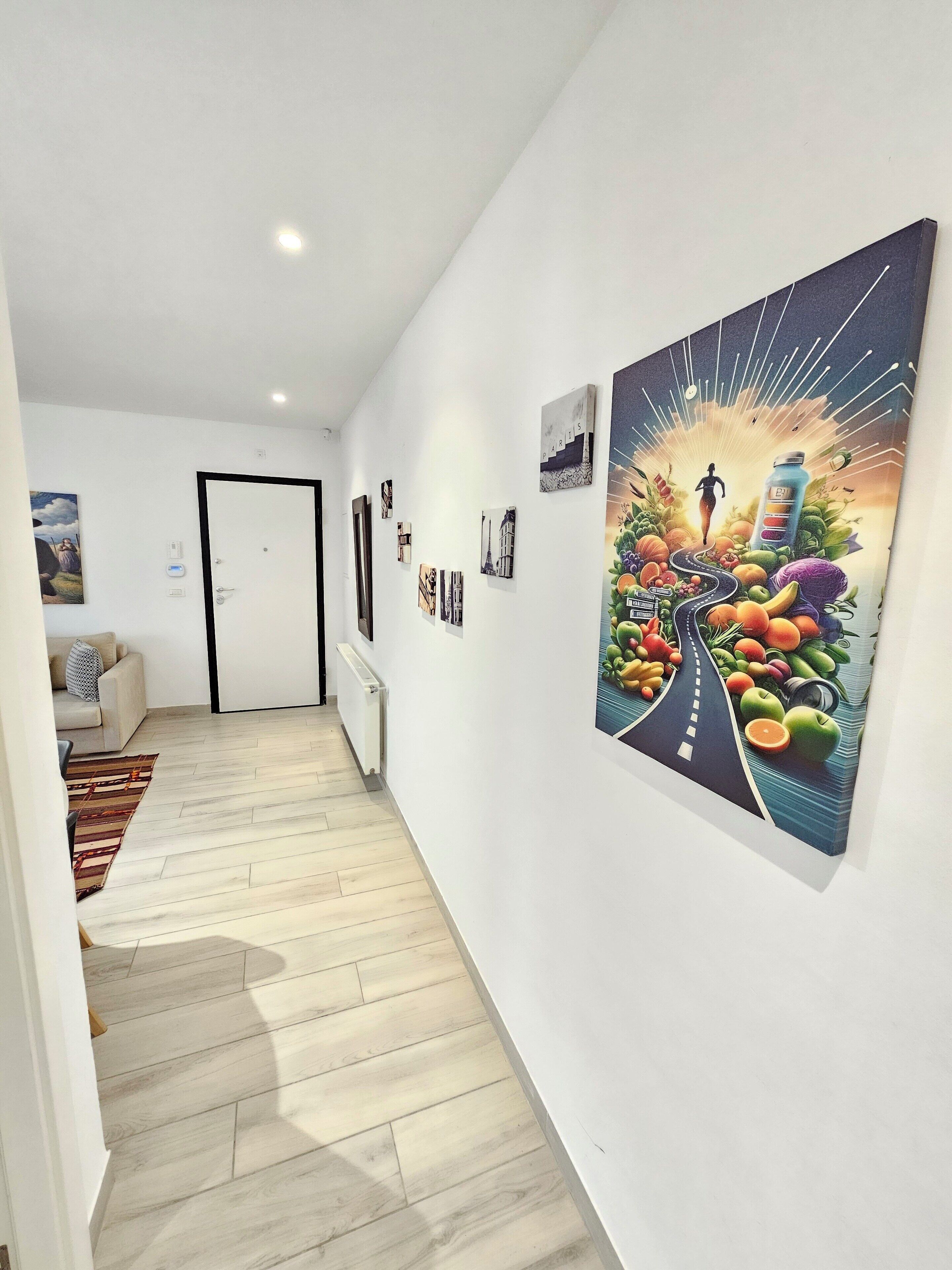 A hallway with a wooden floor, white walls, and a door at the end. There are several framed pictures on the walls, including one of a cityscape and another of a road with fruits and vegetables.