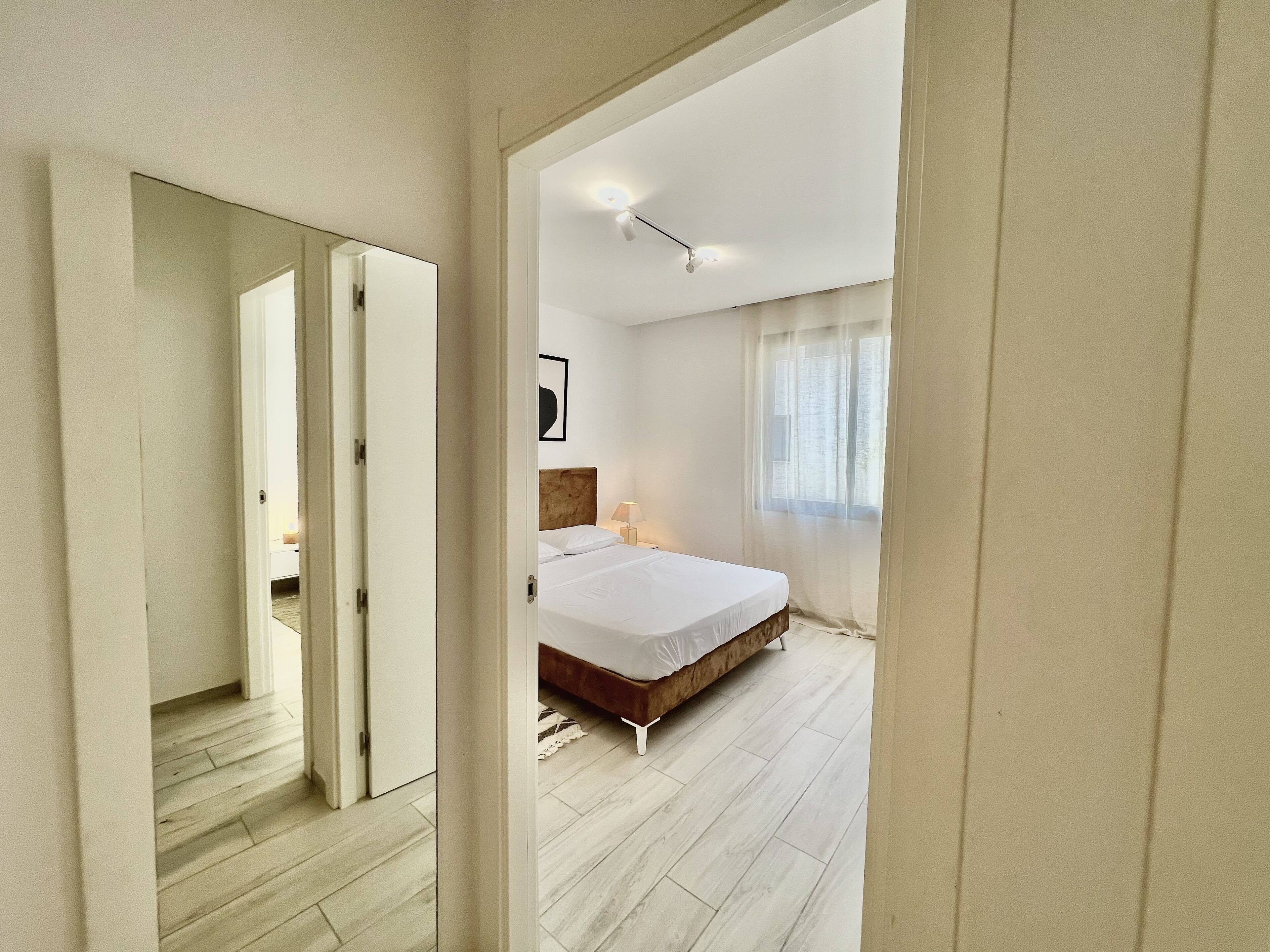 A bedroom with a bed, a nightstand, a window with sheer curtains, and a door leading to another room.