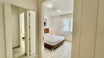 A bedroom with a bed, a nightstand, a window with sheer curtains, and a door leading to another room.