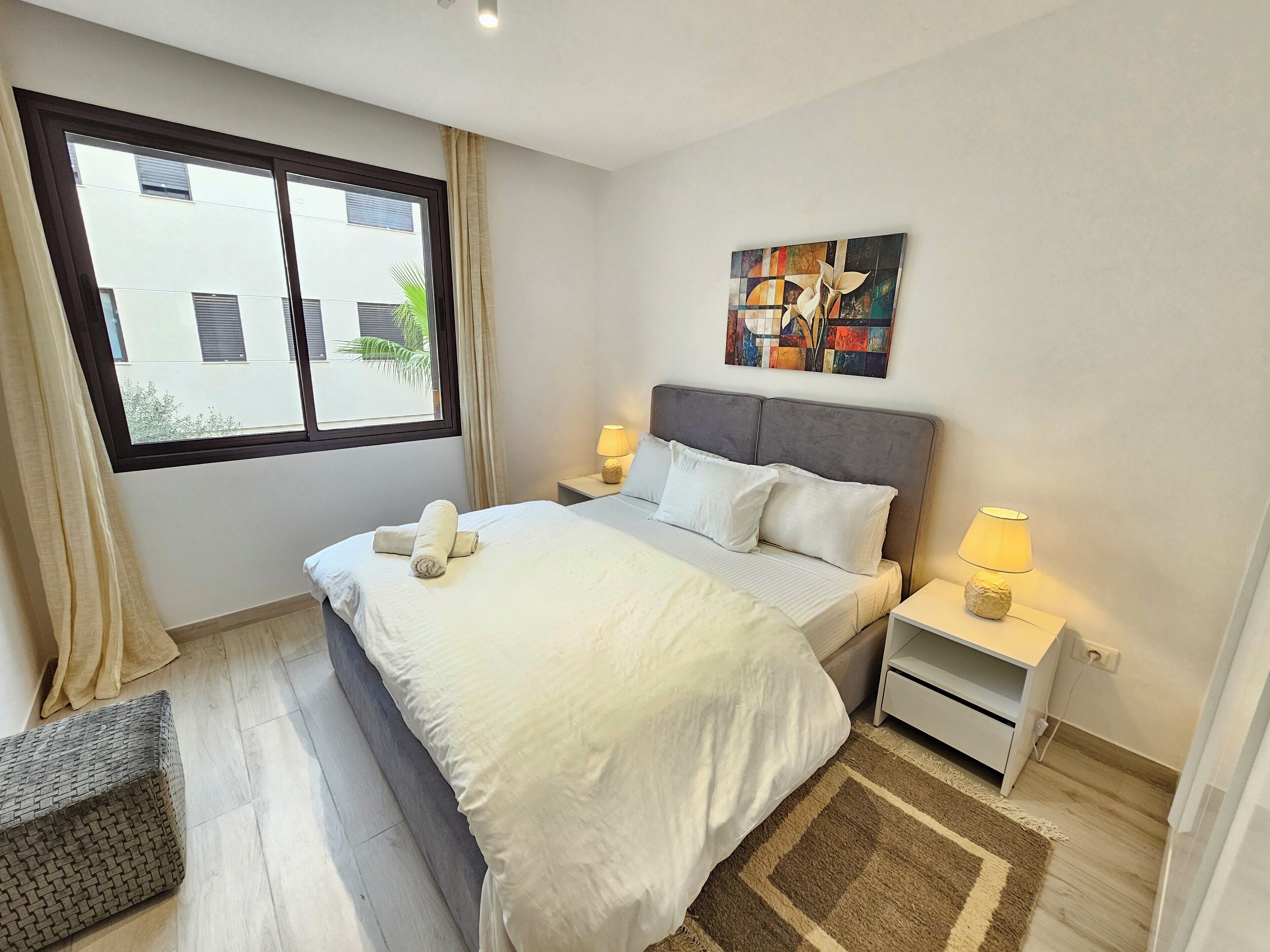 A neatly arranged bedroom with a large bed, two bedside tables with lamps, a window with curtains, and a colorful abstract painting on the wall.
