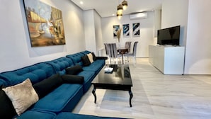 Een moderne woonkamer met een blauwe bank, een eettafel en stoelen. Er is een televisie en een schilderij aan de muur.