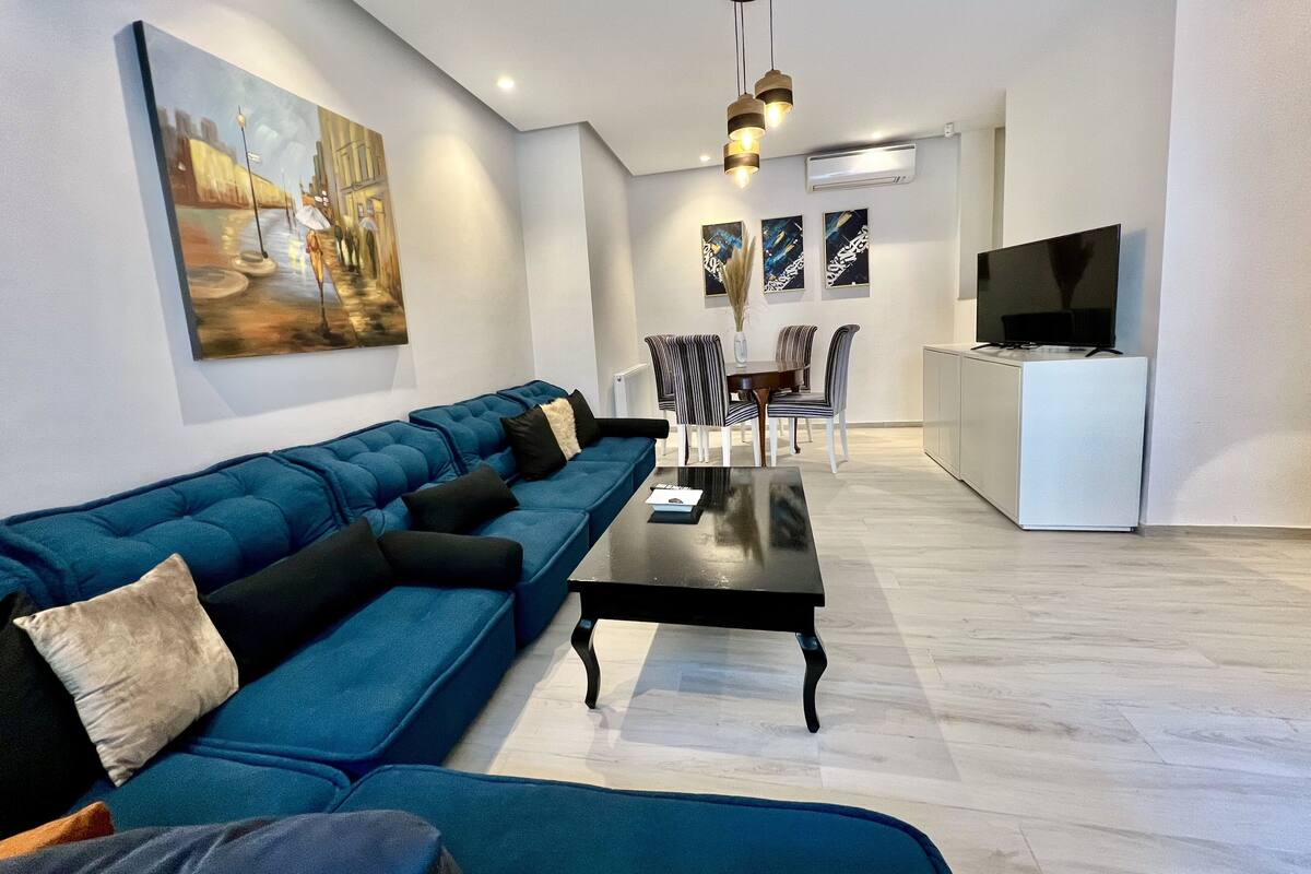 Ein modernes Wohnzimmer mit einem blauen Sofa, einem Esstisch und Stühlen. An der Wand befinden sich ein Fernseher und ein Gemälde.