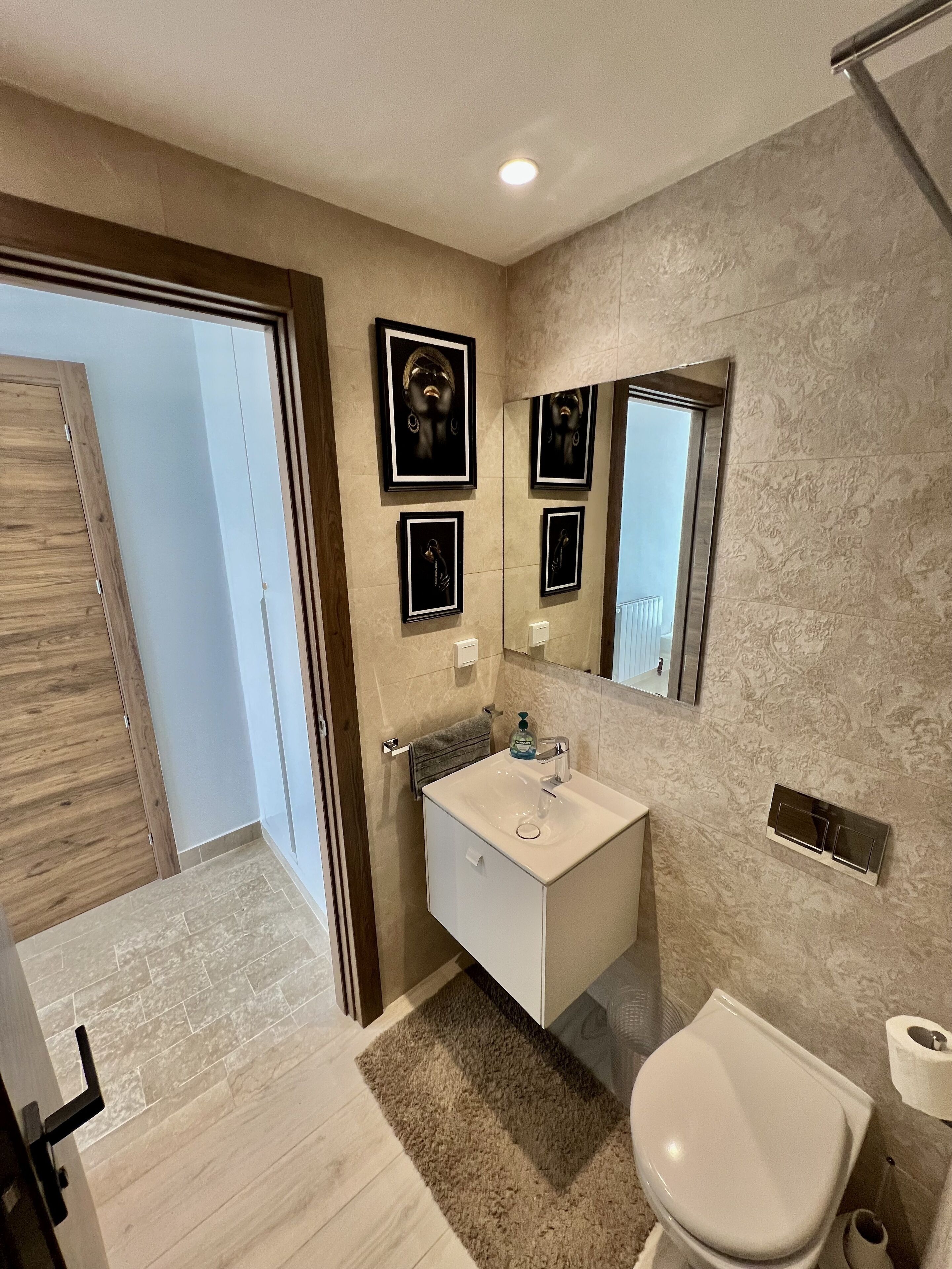 A modern bathroom with a white sink, mirror, and toilet. There are framed pictures on the wall and a wooden door.