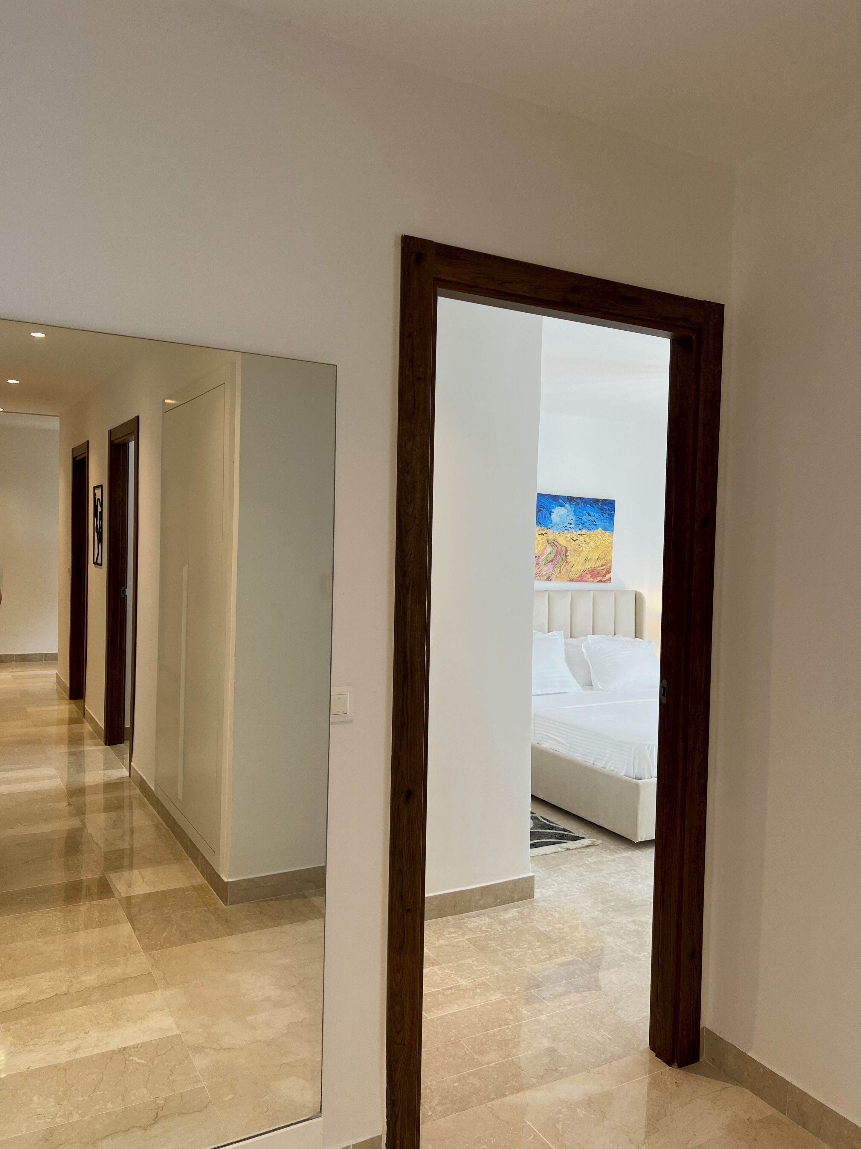 A hotel hallway with a bed in the background, a painting on the wall, and a wooden door frame.