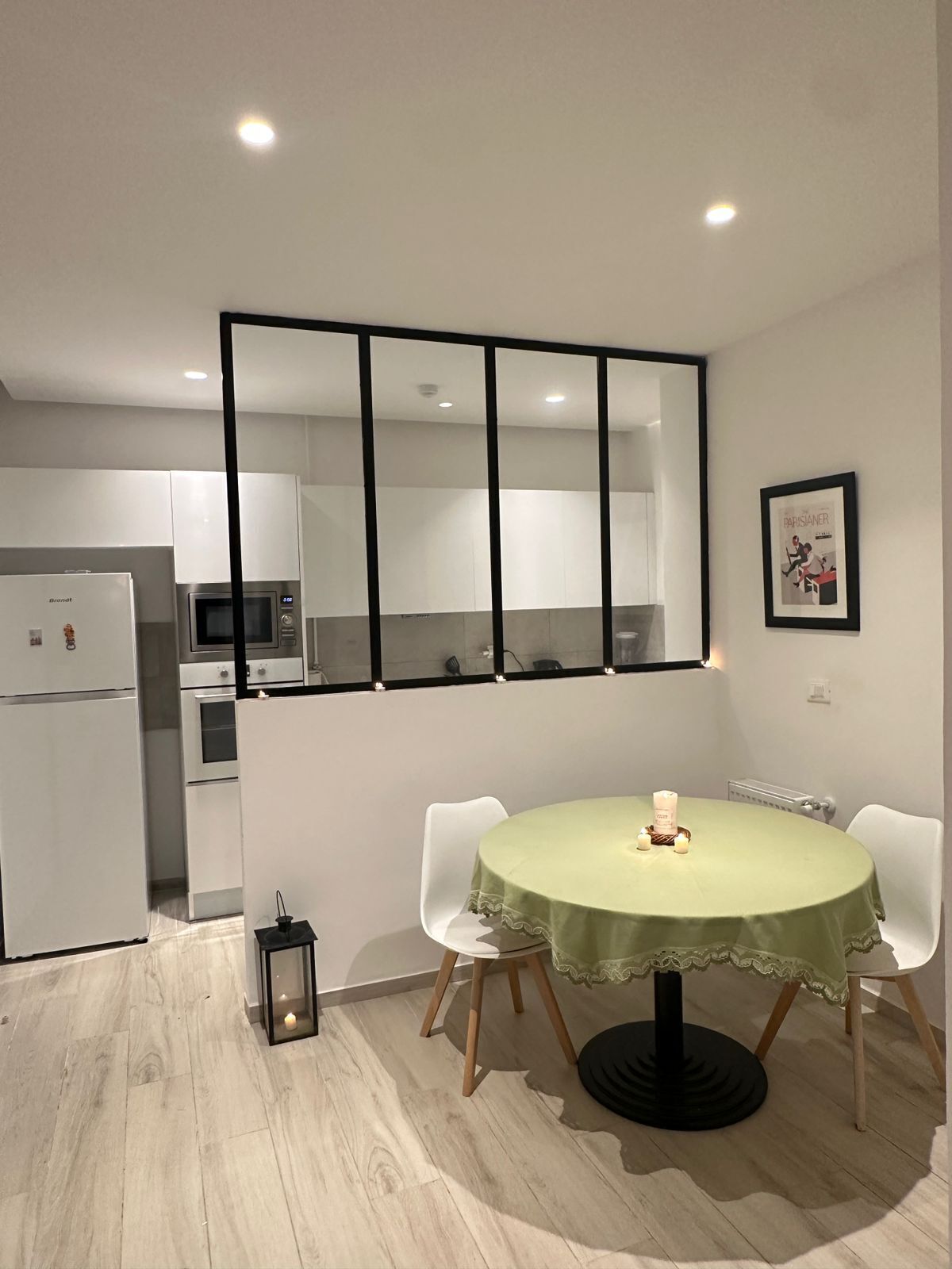 A modern kitchen with white cabinets and a built-in oven. There is a dining area with a round table covered with a green tablecloth and two white chairs. A framed picture hangs on the wall above the table.