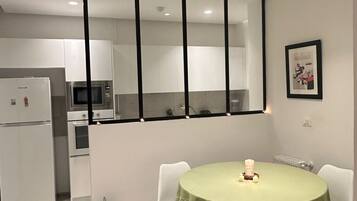 A modern kitchen with white cabinets and a built-in oven. There is a dining area with a round table covered with a green tablecloth and two white chairs. A framed picture hangs on the wall above the table.
