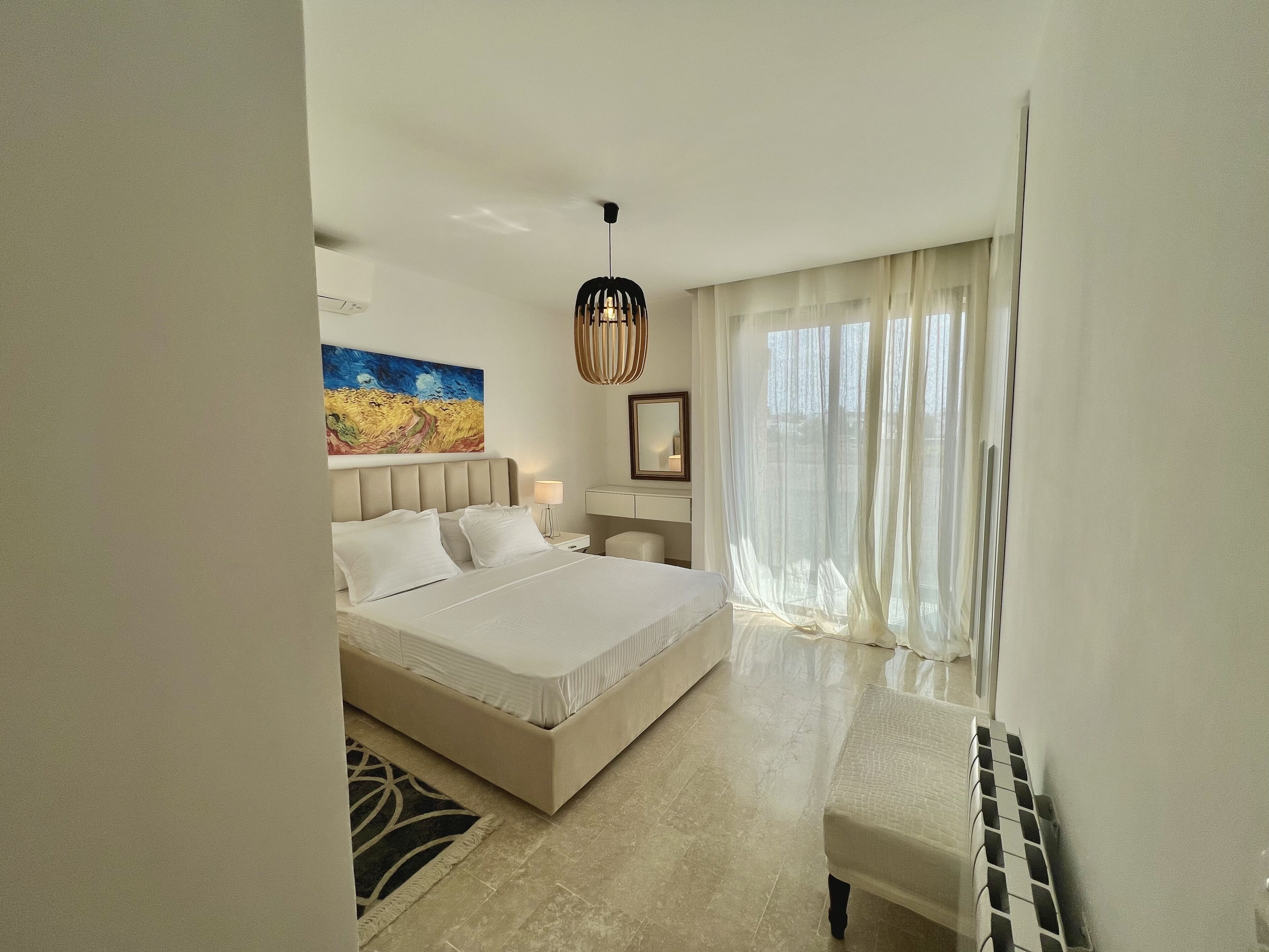 Une chambre d’hôtel avec un grand lit, une table de chevet, un bureau, une chaise, une fenêtre avec des rideaux légers et un tableau au mur.