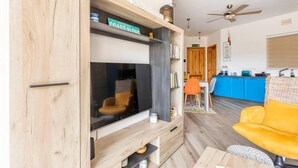 A living room with a wooden entertainment unit, a yellow sofa, and a kitchen area in the background. - Gozo Escape, 2-bed Apartment in Marsalforn (Zebbug)