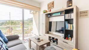 A modern living room with a large TV, bookshelves, and a balcony with a view. - Gozo Escape, 2-bed Apartment in Marsalforn (Zebbug)