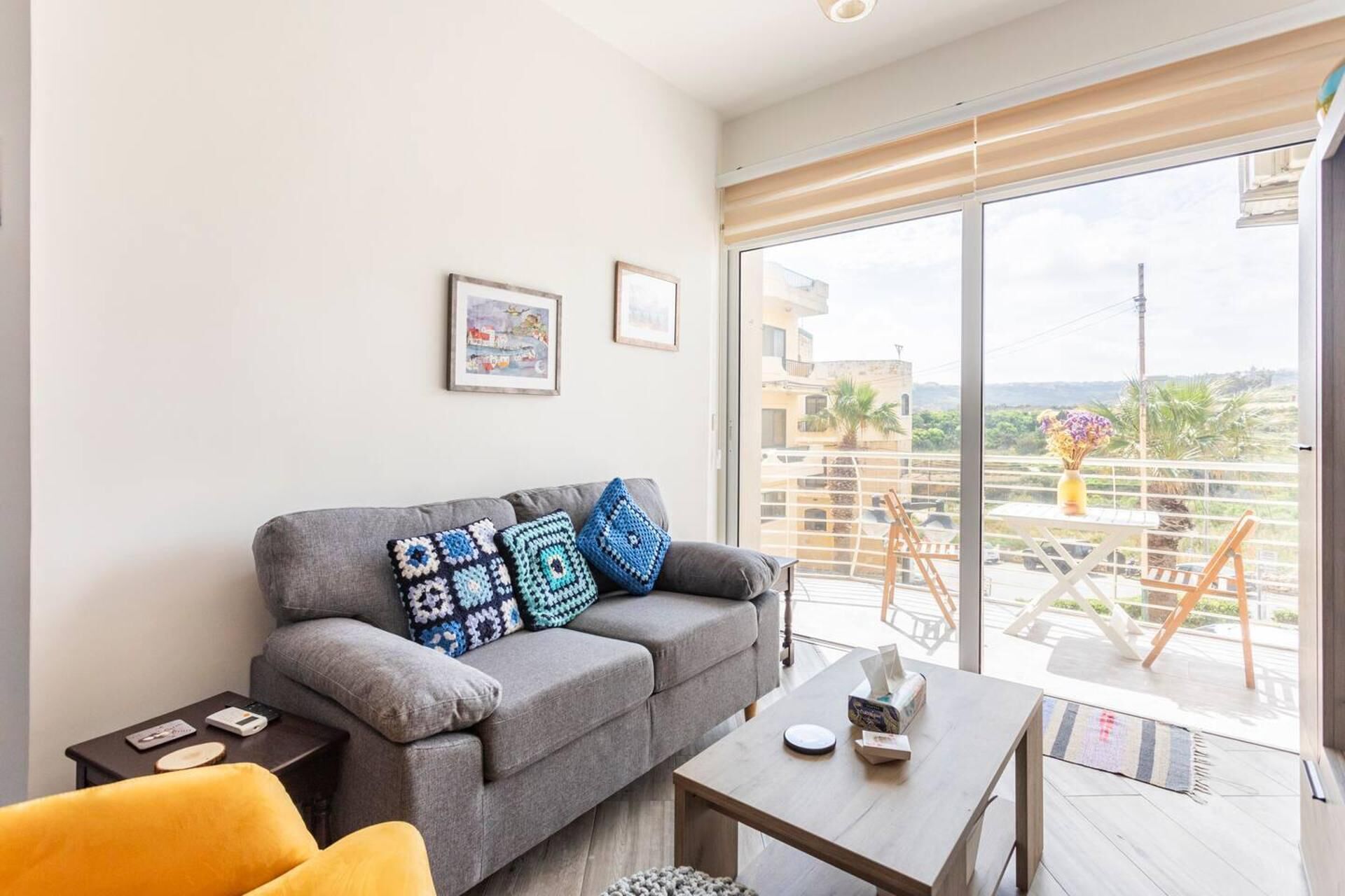 A modern living room with a grey sofa, a yellow armchair, and a wooden coffee table. There are two framed pictures on the wall and a balcony with a view of the city.