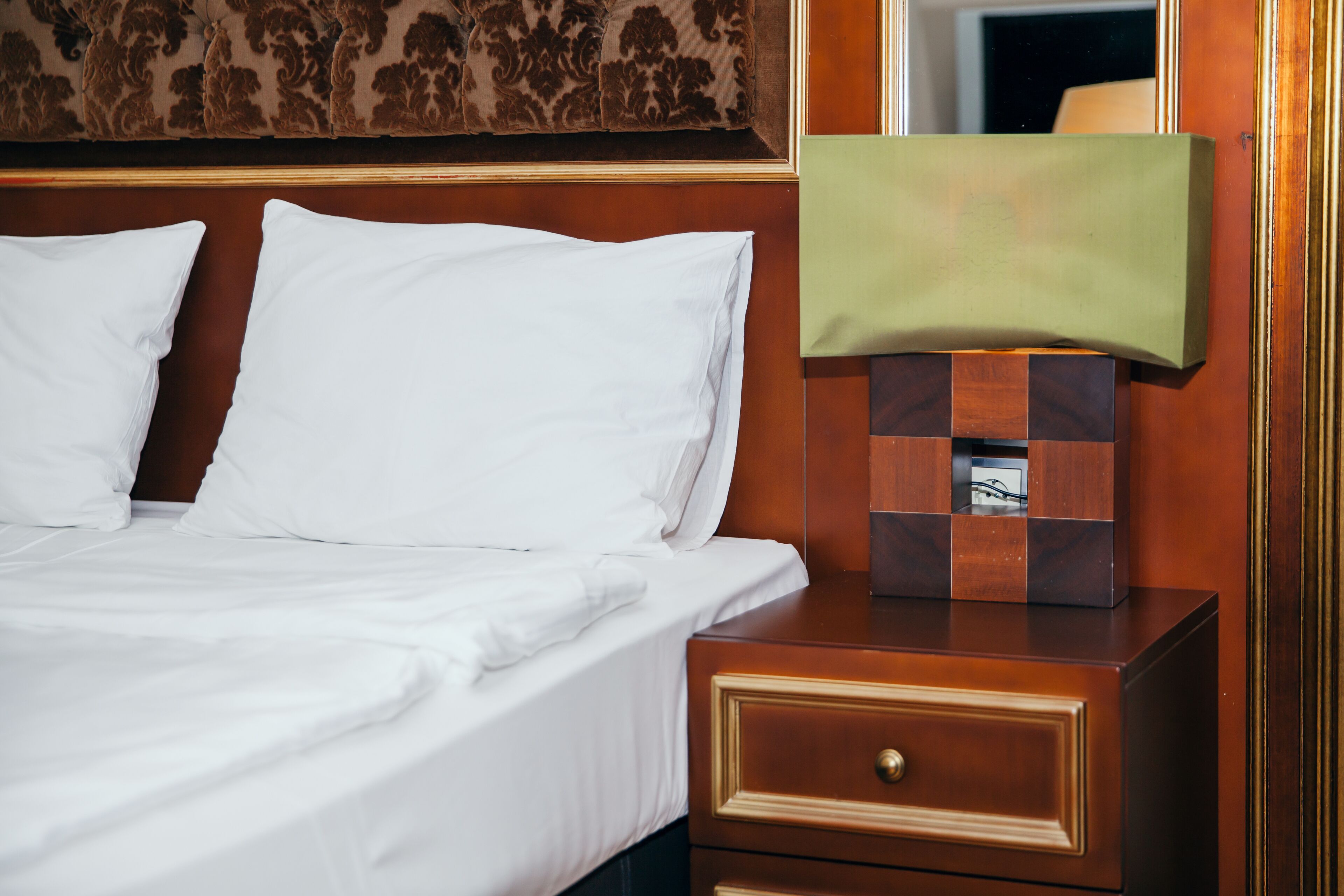 Una cama bien tendida con sábanas blancas, una mesita de noche de madera con cajón y un espejo con una pantalla de lámpara verde.