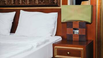 Una cama bien tendida con sábanas blancas, una mesita de noche de madera con cajón y un espejo con una pantalla de lámpara verde.