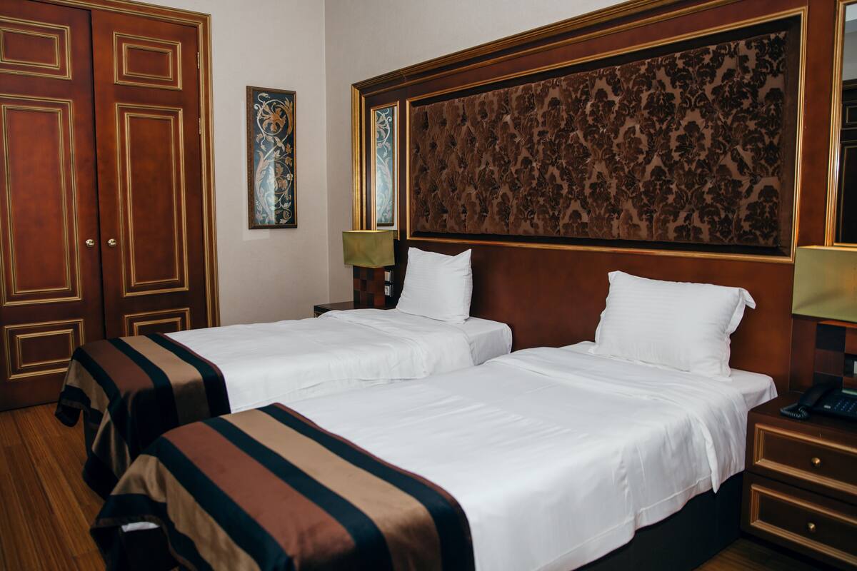 A hotel room with two beds, wooden floors, a large decorative headboard, and a nightstand with a lamp.