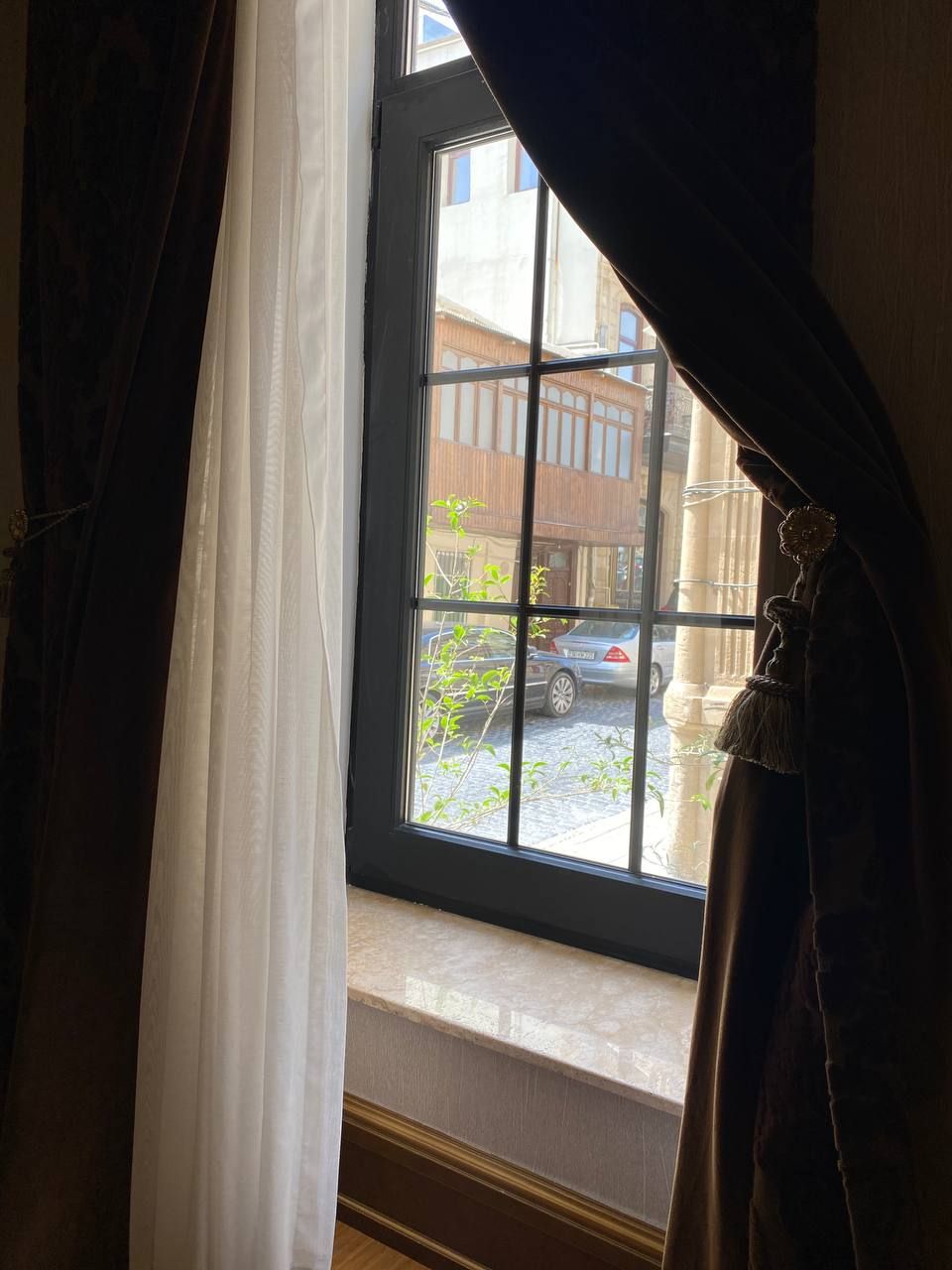 A window with a view of a street, curtains partially drawn, and a glimpse of a building outside.