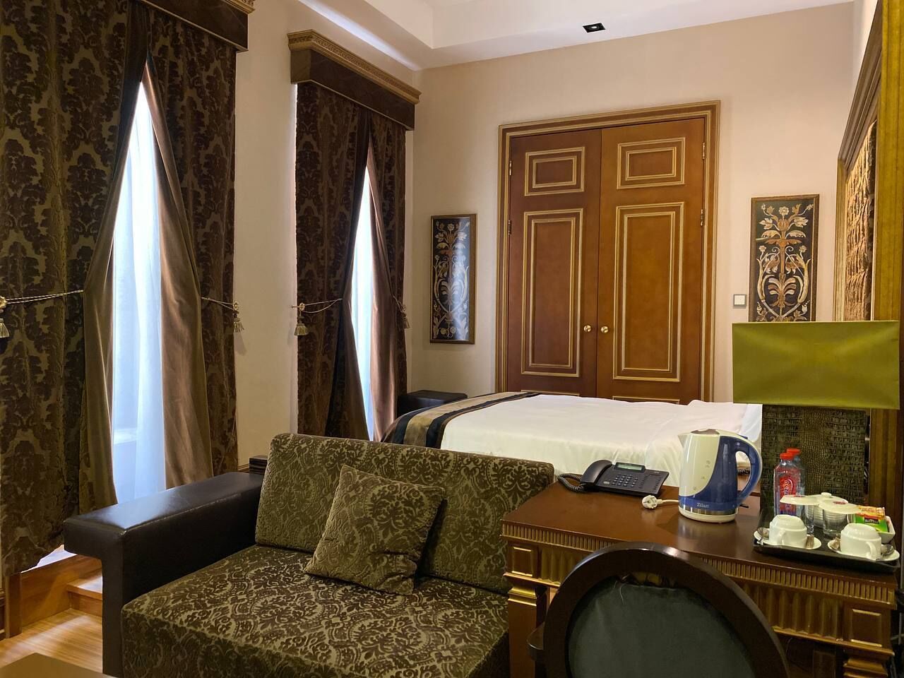A hotel room with a bed, a sofa, a desk with a telephone and a kettle, and a window with curtains.