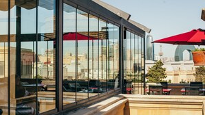 Un área de estar moderna al aire libre con paredes de vidrio, sombrillas rojas y vistas de la ciudad.
