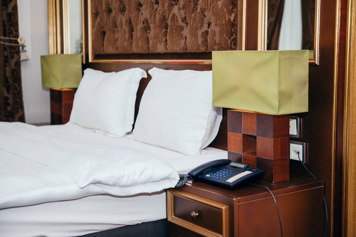 A neatly made bed with white linens, a bedside telephone, and a lamp on a wooden nightstand.