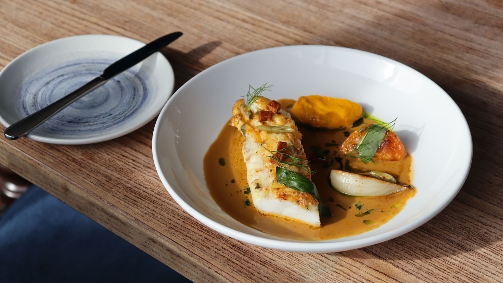 Un plato blanco con un plato de pescado, un bol pequeño con una cuchara y una mesa de madera.