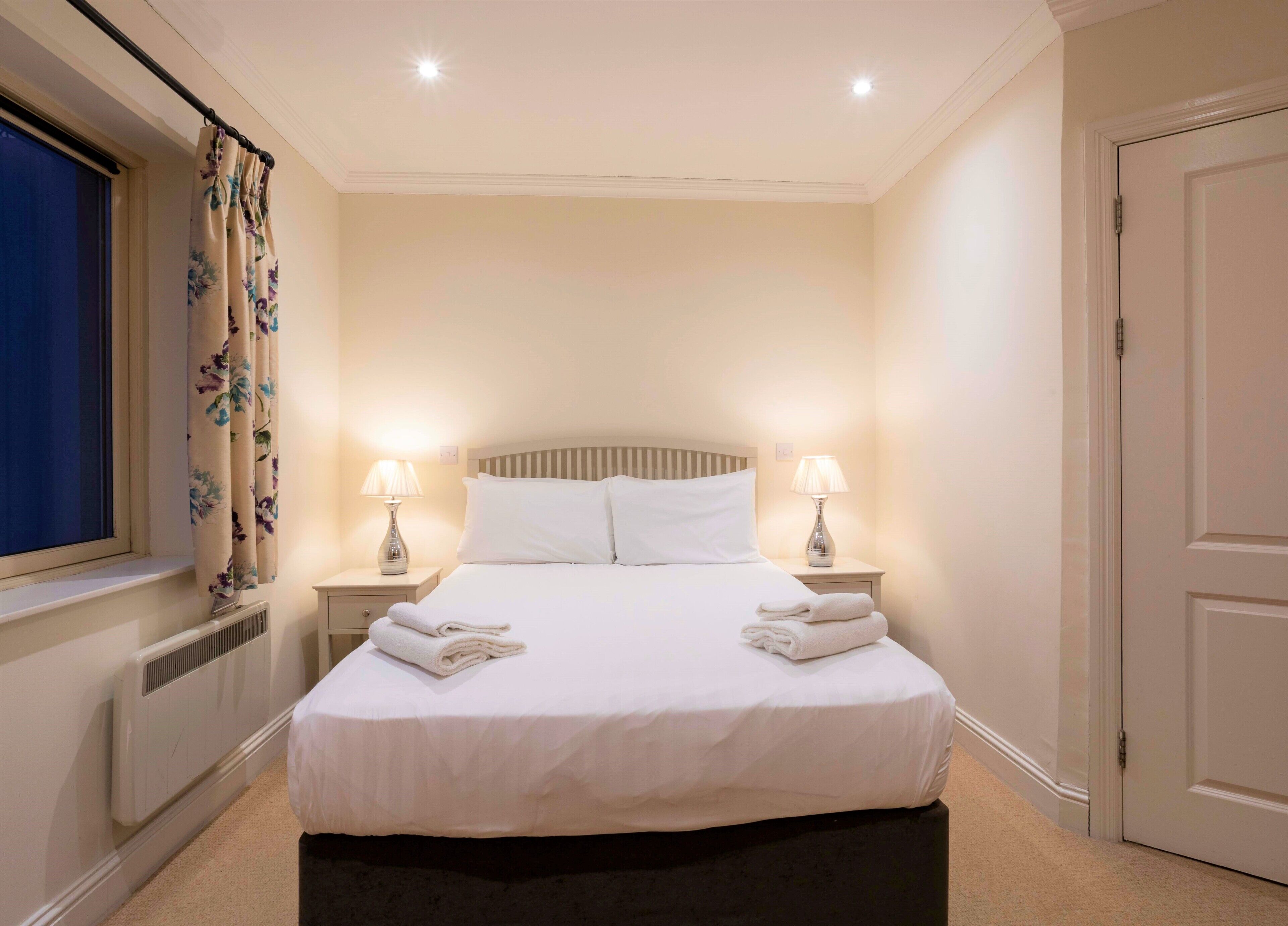 A neatly made bed with white linens, two bedside lamps, and folded towels on the nightstand.