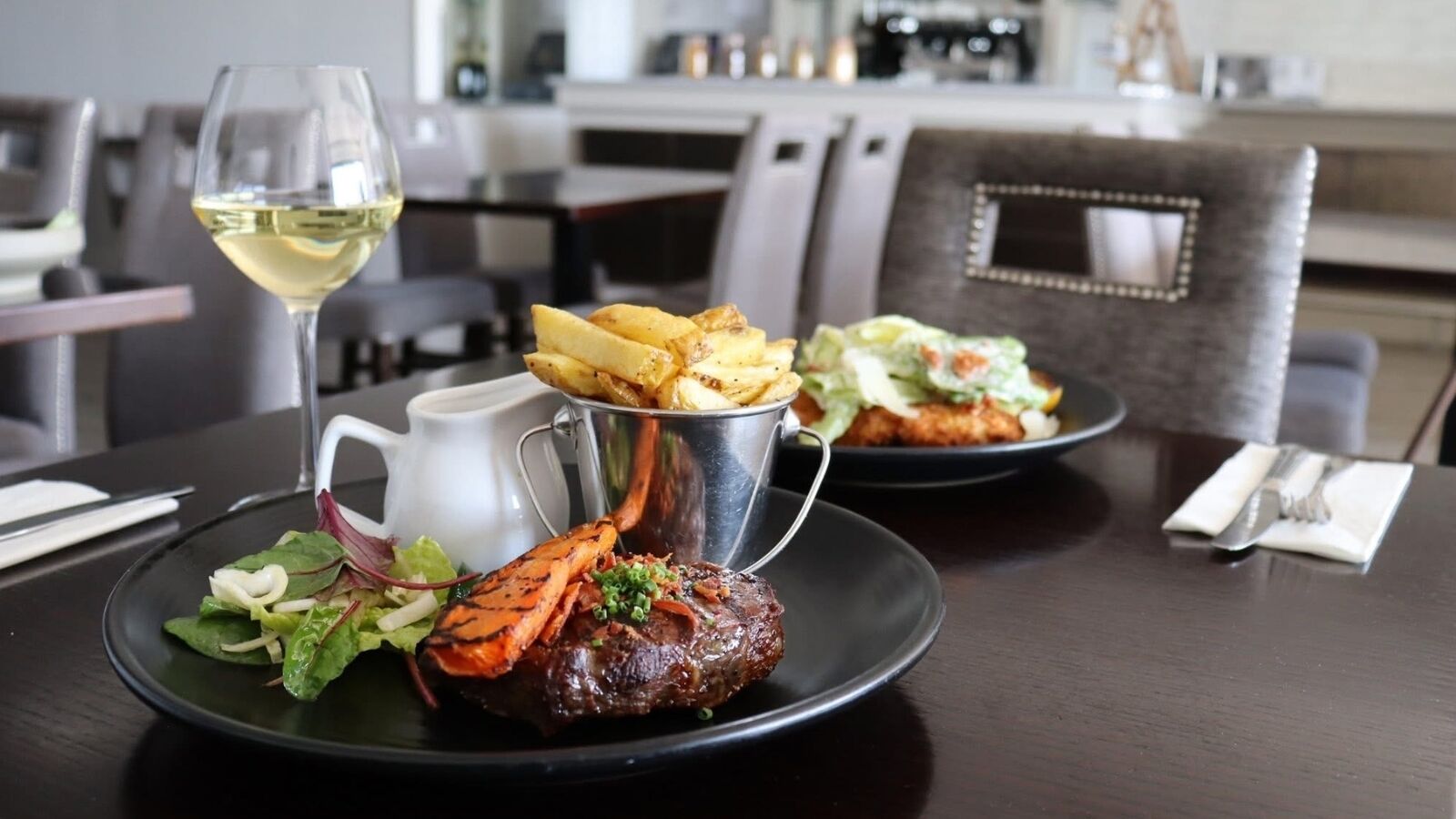 A dining table with a plate of food, a glass of white wine, and a metal pitcher.